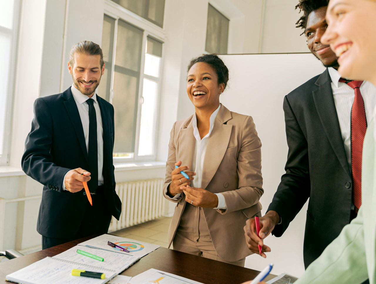 A diverse group of four professionals engaged in a collaborative meeting, smiling and discussing ideas around a table with notebooks and colorful pens.